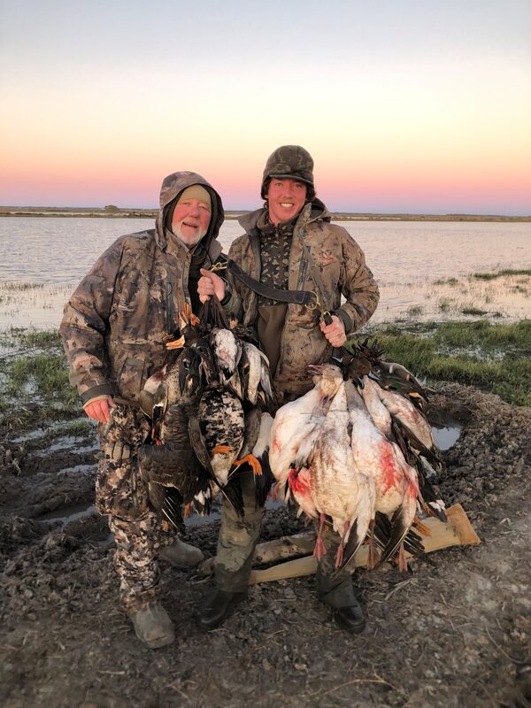 Two hunters standing on a flooded field at first light, holding up the geese they took, with open water and a pink dawn sky behind them.